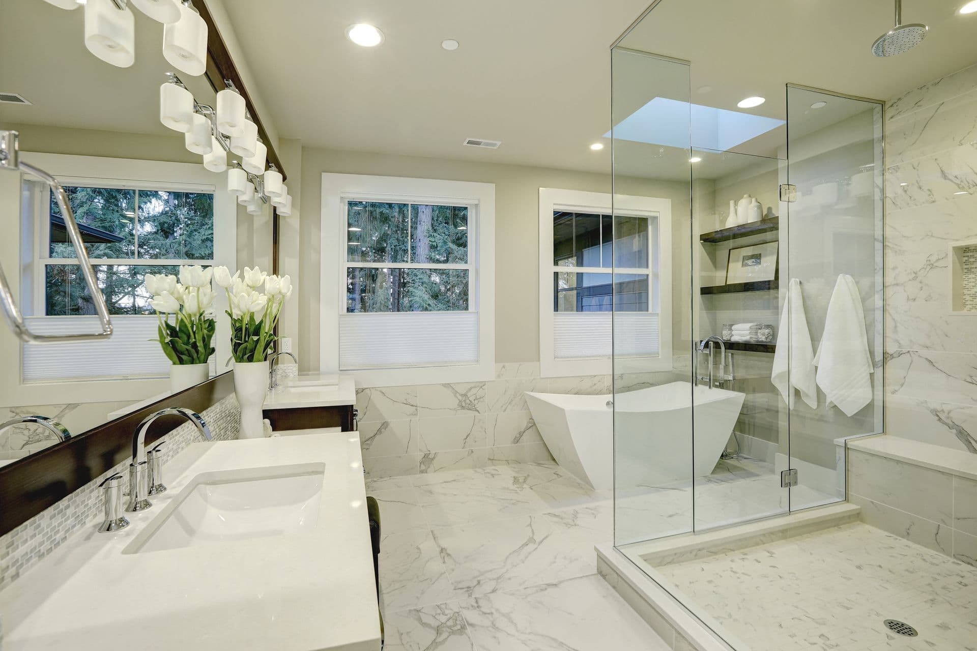 Modern bathroom with marble walls and floor, a glass shower, freestanding tub, large windows, double vanity, and white tulips in a vase; elegant and serene.