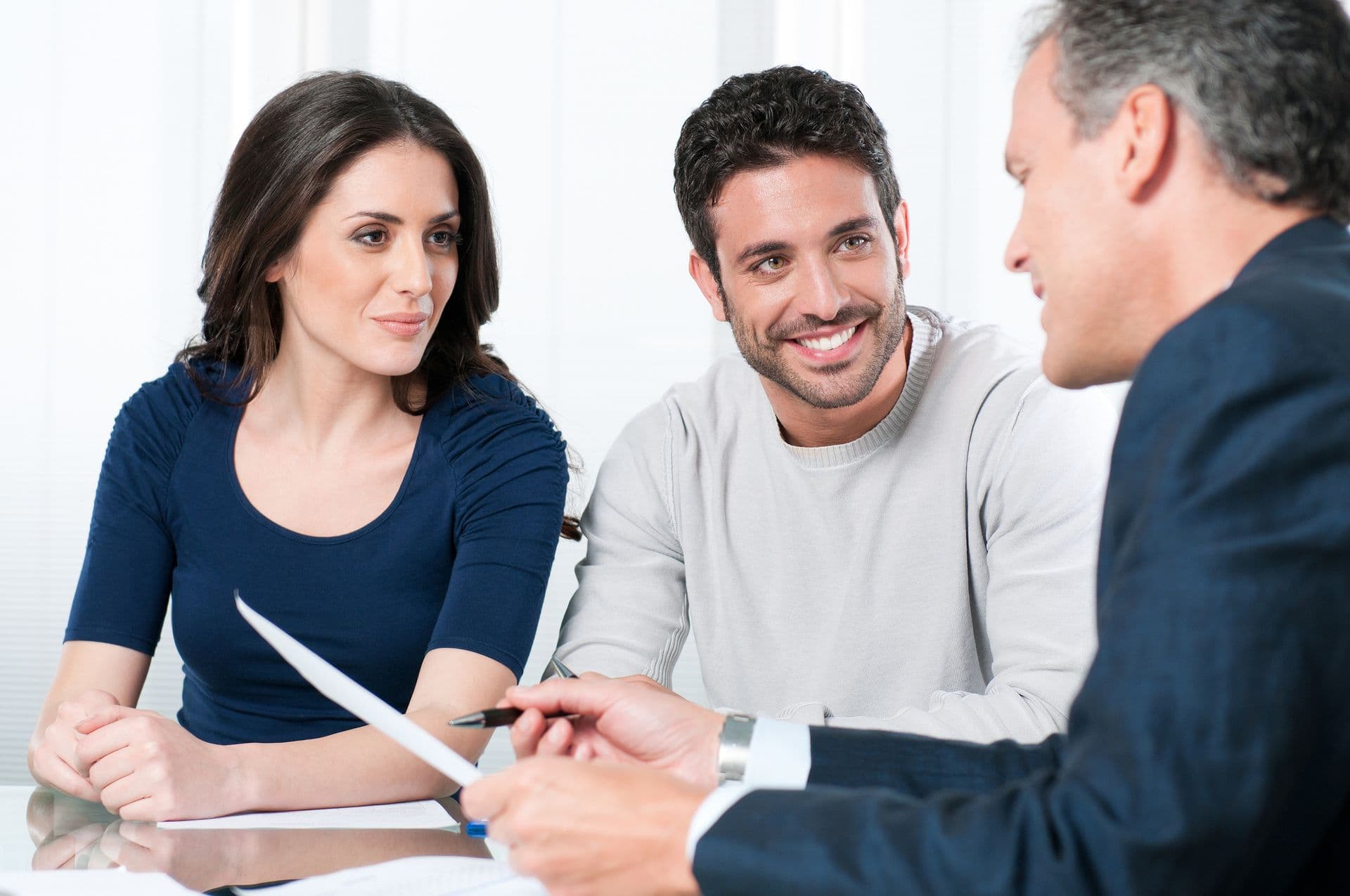 A couple sits smiling across a desk from a suited man holding documents. The setting is professional, conveying a positive, cooperative atmosphere.