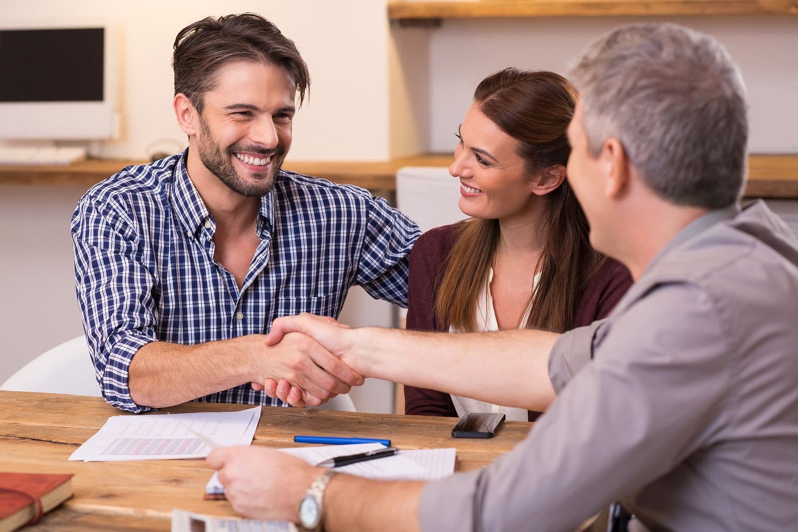 A smiling man in a plaid shirt shakes hands with another man across a table, while a woman beside him smiles. Papers and a pen are on the table.