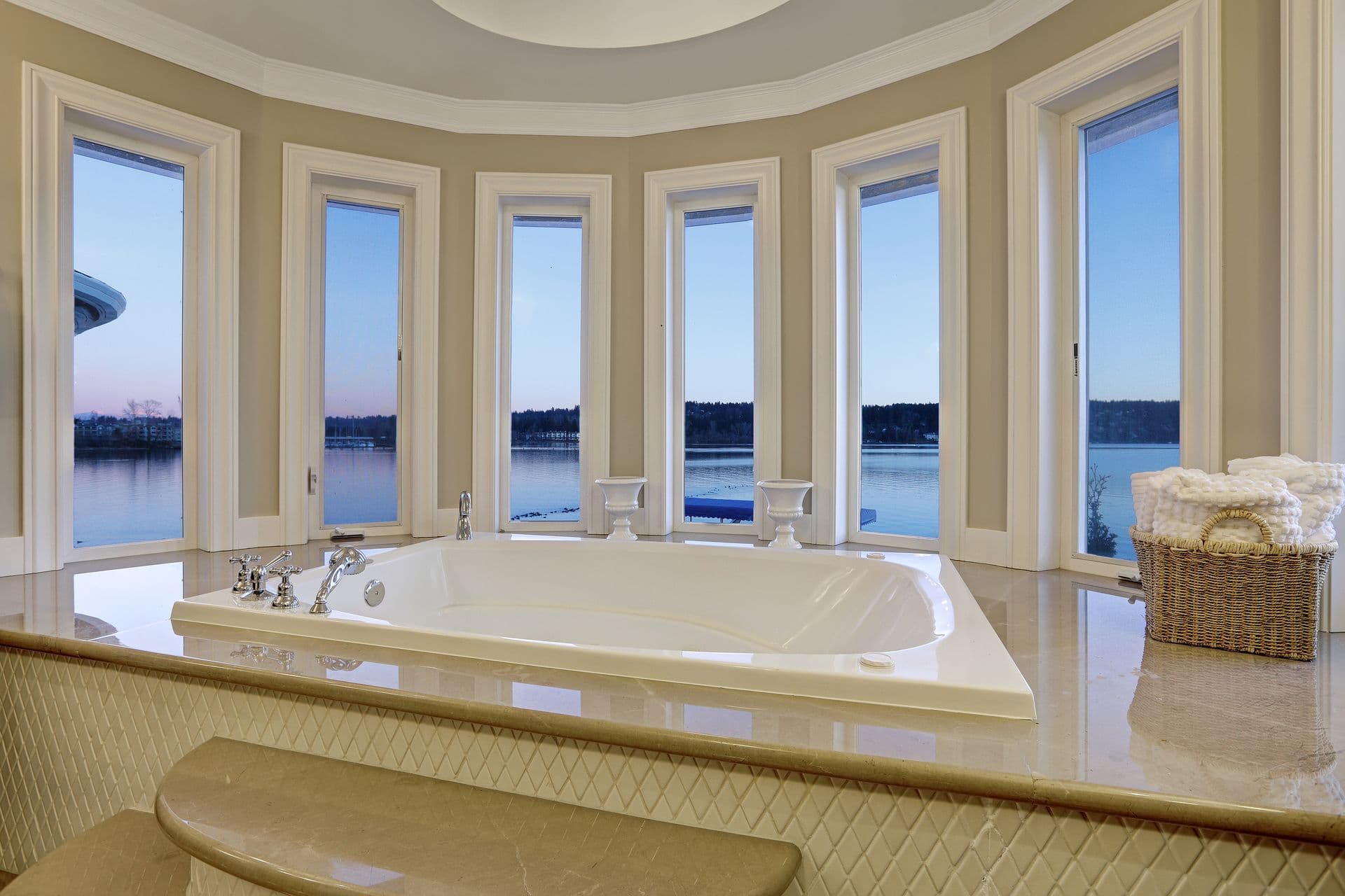 Luxurious bathroom with a large white bathtub surrounded by tall windows, overlooking a serene lake view at dusk. A basket of towels adds warmth.