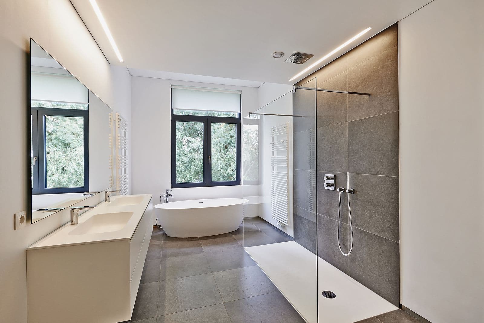Modern bathroom with sleek design. Features include a freestanding bathtub, large mirror, dual sinks, glass-enclosed shower, and a window with a garden view.