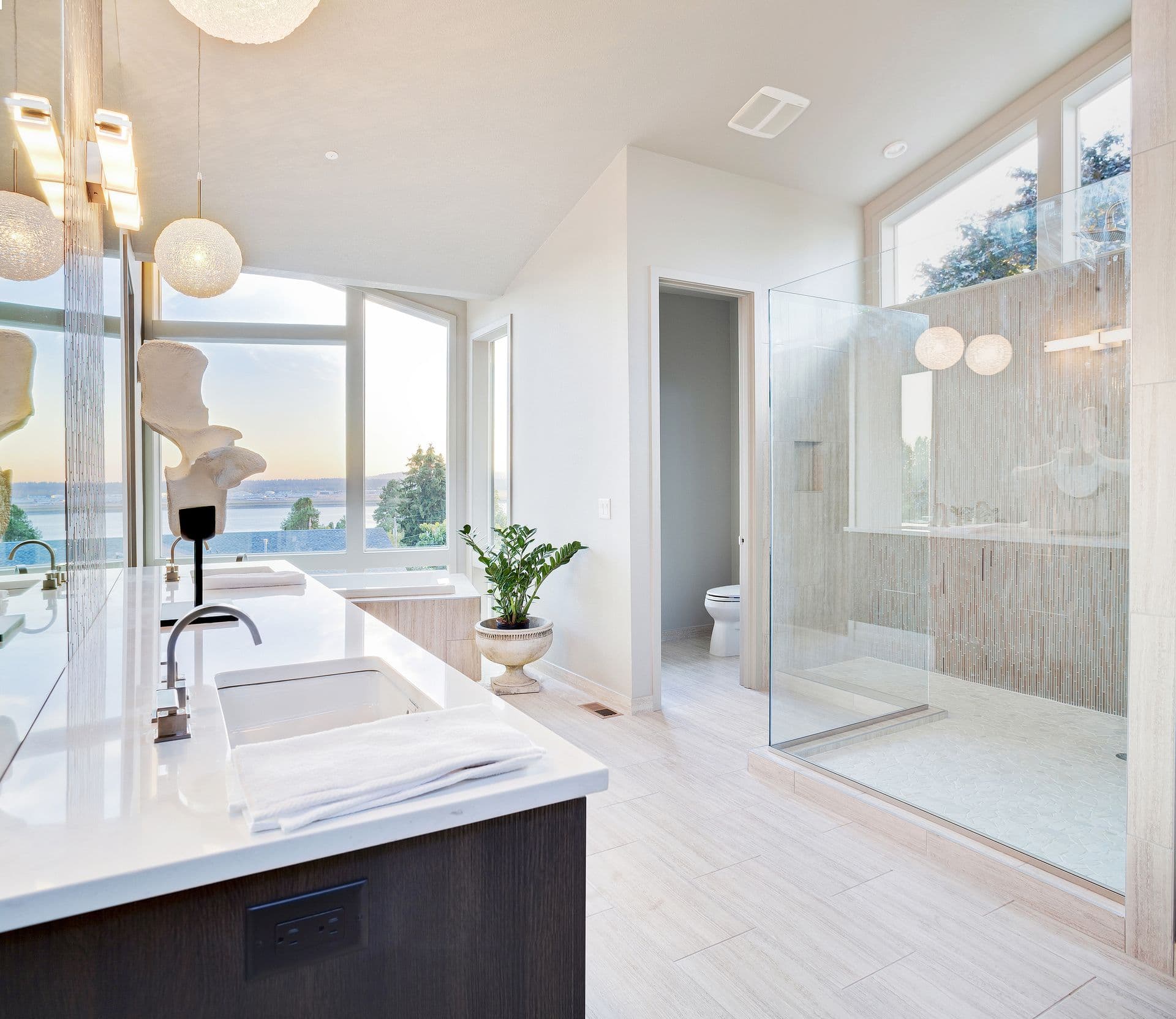 Modern bathroom with large windows showing a scenic view. Features a double vanity with a white countertop, glass shower, and potted plant. Bright and serene ambiance.