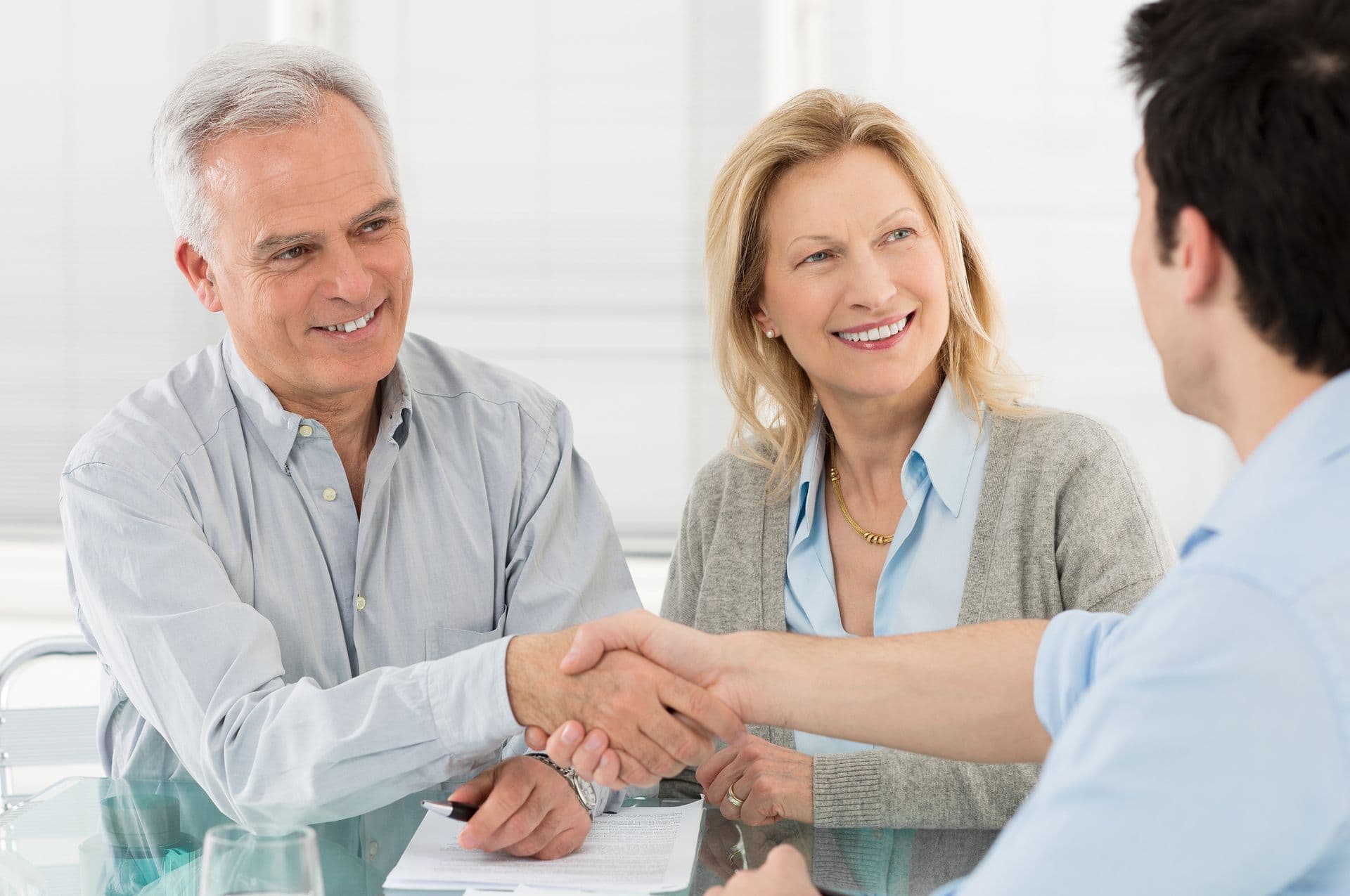 Older couple smiling as they shake hands with a younger man across a glass table, conveying a sense of agreement and positive communication.