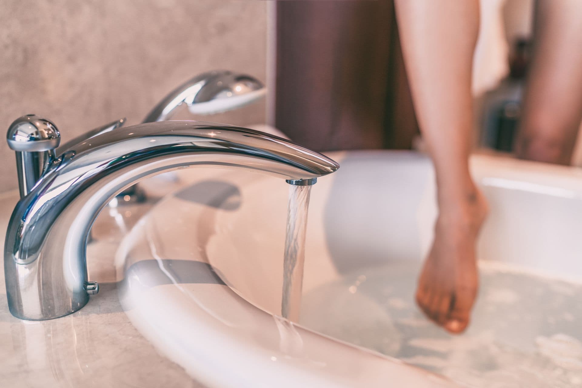 A chrome faucet pours water into a bathtub, while a person's foot steps in, suggesting a relaxing bath. The scene feels calm and inviting.