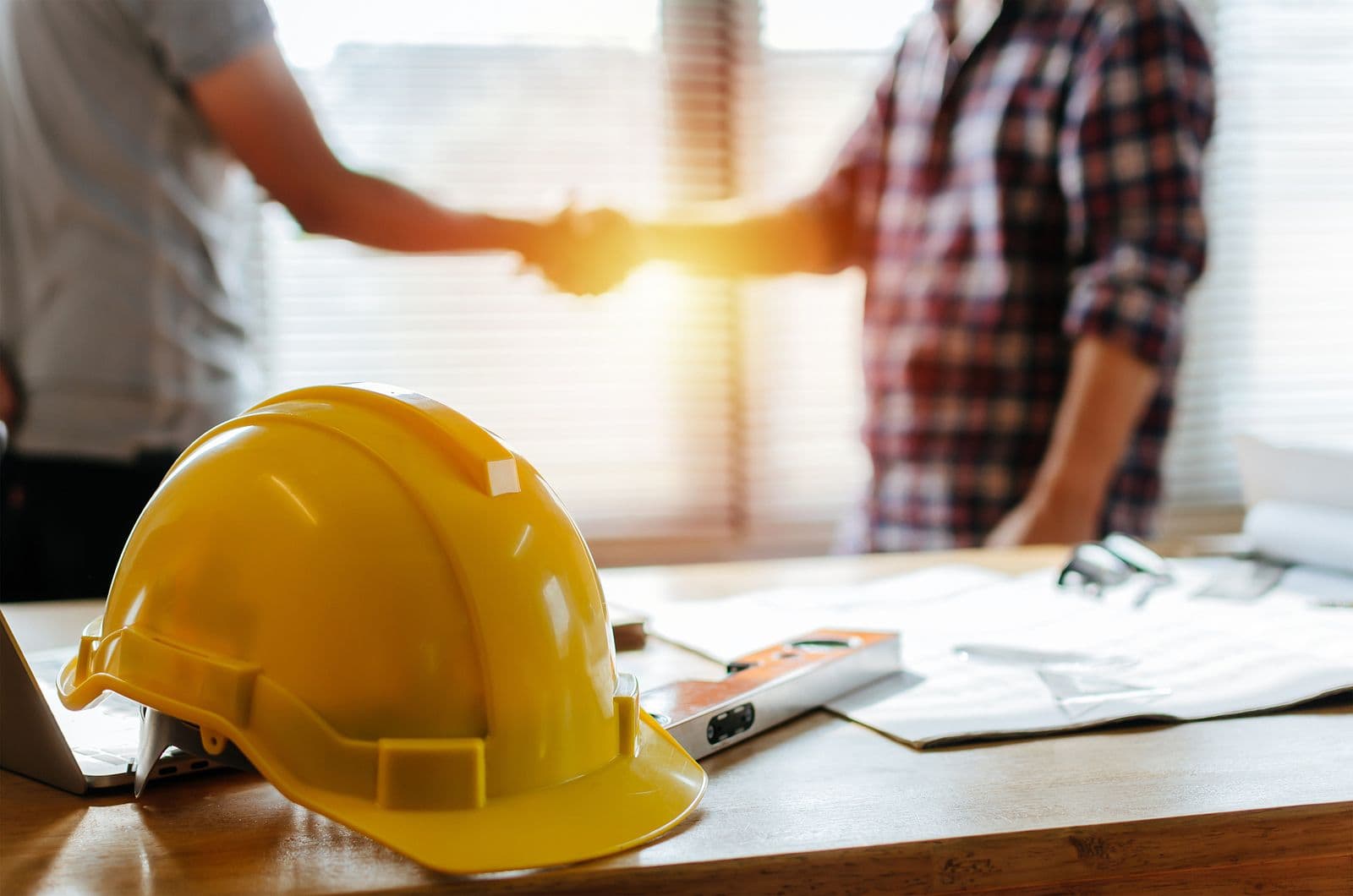 Yellow hard hat on a desk with blueprints, a level, and a laptop. In the background, two people shake hands, conveying agreement and teamwork.