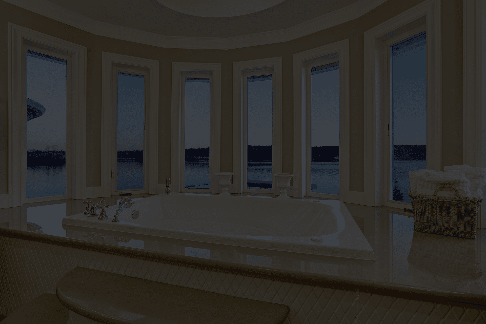 Luxurious bathroom with a large, white bathtub surrounded by tall windows showing a serene lake view at sunset. There’s a basket with towels nearby.