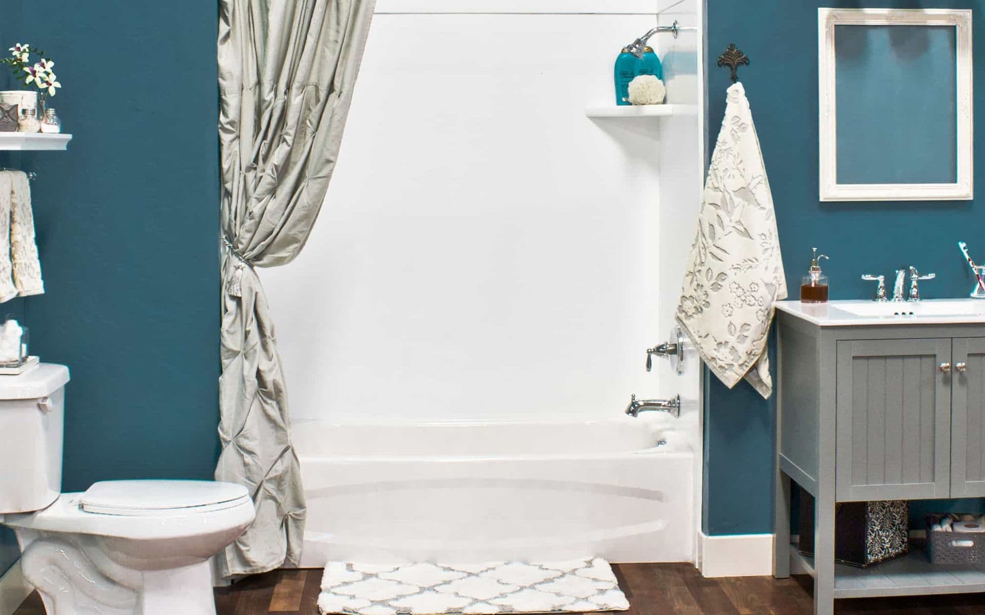 A cozy bathroom with blue walls features a white bathtub with a curtain, a modern gray vanity with a mirror, a toilet, and decorative shelves. Warm and inviting.